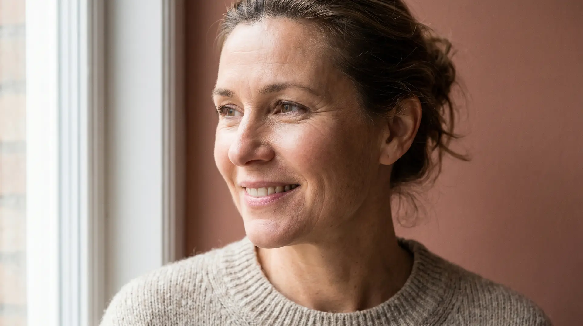 Woman in her late 40s with healthy luminous skin and a warm smile in soft window light